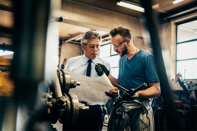Chef und Mitarbeiter besprechen Plan und Werkstück in der Werkstatt