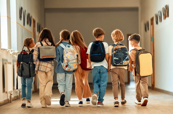 7 Kinder mit Rucksäcken gehen gemeinsam Gang eines Schulgebäudes entlang