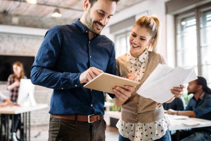 In einem hellen, modernen Büro unterhalten sich zwei Personen, während sie Dokumente und ein Tablet durchsehen. Der Arbeitsplatz zeichnet sich durch große Fenster, freigelegte Backsteinwände und eine ungezwungene, aber dennoch professionelle Atmosphäre aus. Im Hintergrund sind weitere Teammitglieder zu sehen, die zu einem kooperativen Arbeitsumfeld beitragen.