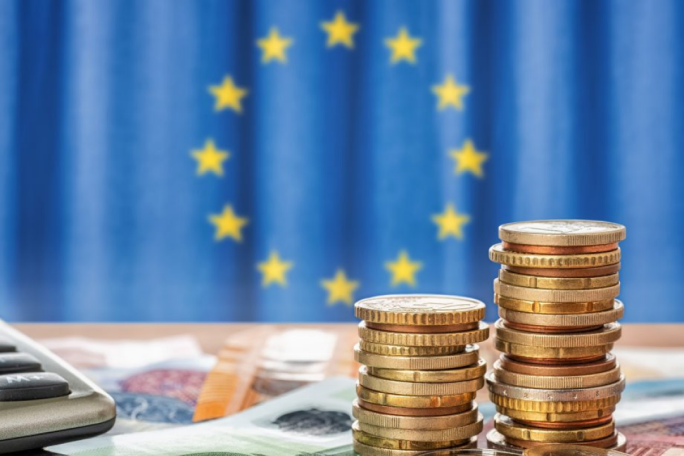 Stacks of Euro coins are placed on a table alongside a calculator and Euro banknotes. The European Union flag with yellow stars on a blue background is prominently visible in the background. The image conveys themes of finance, economy, and European monetary systems.