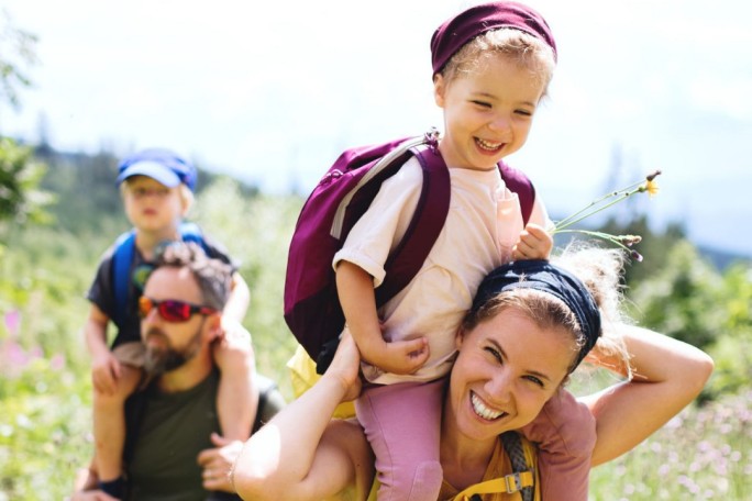 A group of individuals enjoying a hike in a lush, mountainous setting. The scene features adults and children wearing backpacks, with one child holding a small flower. Bright daylight and greenery dominate the visuals, creating a vibrant and natural atmosphere. The activity suggests outdoor exploration and family bonding.