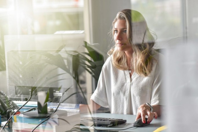 A woman is seated at a desk in a modern office environment, using a computer mouse and keyboard. The setting features natural light streaming through large windows, with indoor plants adding a touch of greenery. Office supplies and technology are visible, creating a professional workspace atmosphere.