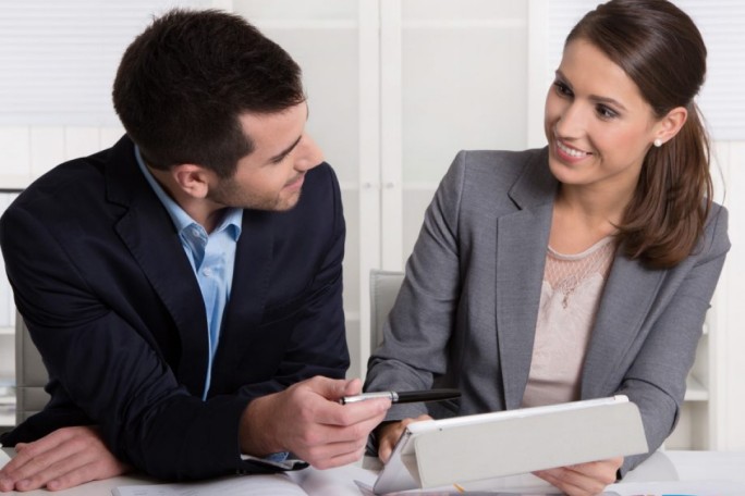 Two business professionals are seated at a desk in a modern office setting, engaged in a discussion while using a tablet device. The man is holding a pen, pointing towards the tablet, while the woman attentively listens. Both are dressed in formal business attire, suggesting a professional environment.