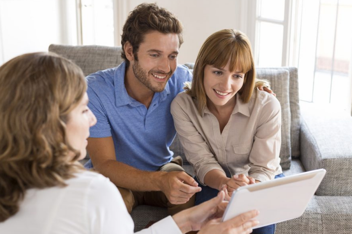 A couple sits on a couch in a bright living room while engaging with an advisor using a tablet. The setting features natural light and a casual atmosphere. The focus is on collaboration and technology in a professional or personal consultation.