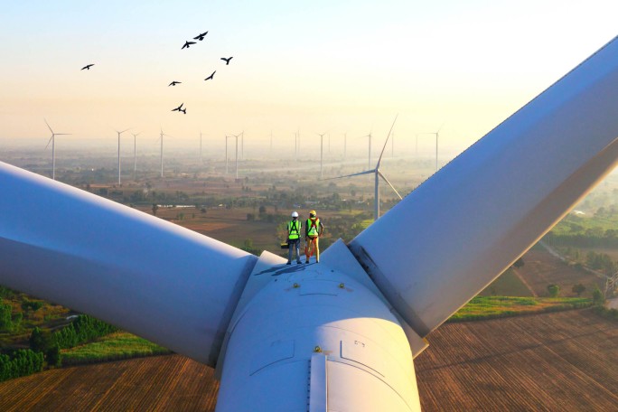 Zwei Arbeiter in Schutzausrüstung stehen auf einer großen Windkraftanlage und überblicken bei Sonnenaufgang ein weitläufiges Windkraftfeld. Vögel ziehen ihre Kreise und verleihen der friedlichen Landschaft eine dynamische Note. Die Szenerie unterstreicht die Bedeutung erneuerbarer Energien und die Instandhaltung von Industrieanlagen im ländlichen Raum.