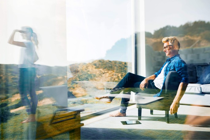 Ein Mann sitzt entspannt in einem modernen Innenraum mit großen Glasfenstern, die einen malerischen Blick auf die Hügel freigeben. Draußen steht eine Frau und betrachtet die Landschaft mit einem Fernglas. Die Atmosphäre ist hell und ruhig und verbindet den Komfort des Innenraums mit der Erkundung der Natur.