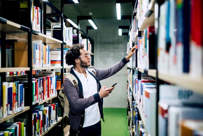 Ein junger Mann mit Rucksack stöbert in einer hellen, modernen Bibliothek. In der einen Hand hält er sein Smartphone, mit der anderen greift er nach einem Buch im Regal. Die Bibliothek besticht durch ordentliche Regale voller bunter Bücher und einen grünen Teppichboden, die eine konzentrierte und zeitgemäße Atmosphäre schaffen.