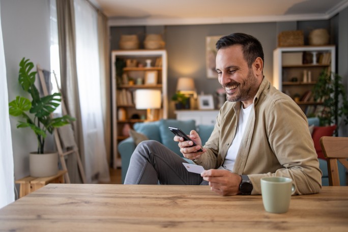 Ein Mann sitzt an einem Holztisch in einem behaglichen Wohnzimmer und hält ein Smartphone und eine Karte in der Hand. Der Raum ist modern eingerichtet mit Bücherregalen, Pflanzen und sanfter Beleuchtung. Eine grüne Keramiktasse steht auf dem Tisch und trägt zur warmen und entspannten Atmosphäre bei.